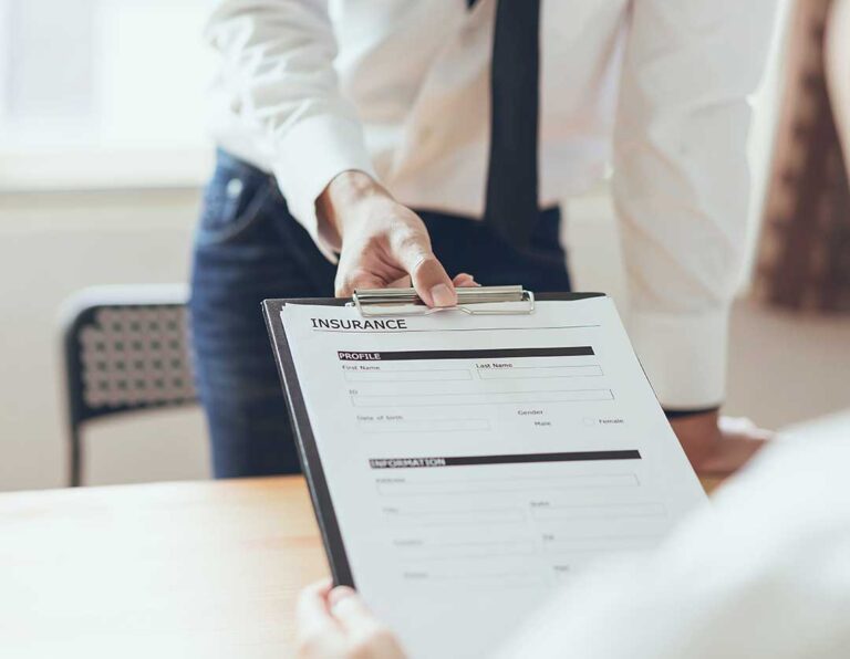 A person sits at a kitchen table reviewing printed insurance policy paperwork with a pen and notebook.